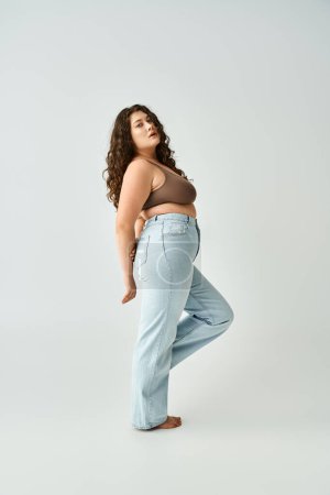 attractive plus size woman in brown bra and blue jeans with hands behind back posing sideways