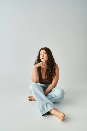 Photo for Attractive plus size young girl in brown bra and blue jeans sitting and touching finger to chin - Royalty Free Image