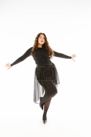 Foto de Hermosa mujer joven con curvas en traje elegante negro con pelo rizado posando con las manos en los lados - Imagen libre de derechos