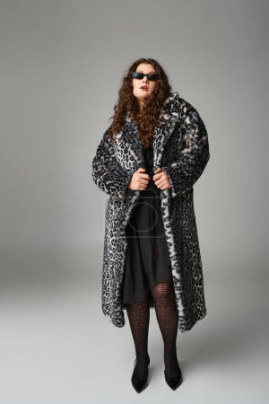 beautiful curvy woman in leopard fur coat and sunglasses standing on grey background