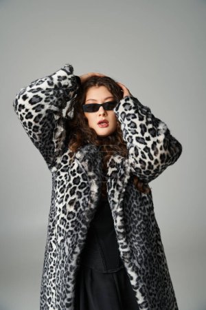 Schöne kurvige Frau in Leopardenfell-Mantel und Sonnenbrille umarmt Kopf mit Händen auf grauem Hintergrund