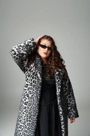 beautiful curvy young woman in leopard fur coat and sunglasses posing with hand on head on grey