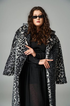 Téléchargez les photos : Femme courbée confiante dans des lunettes de soleil debout avec manteau de fourrure de léopard sur fond gris - en image libre de droit