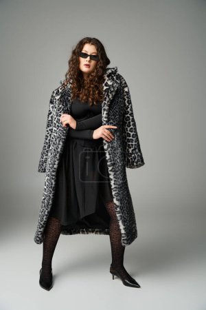 mujer con curvas en gafas de sol de pie con abrigo de piel de leopardo y piernas separadas sobre fondo gris