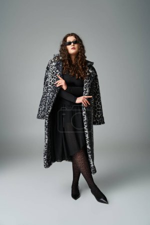 attraktive kurvige junge Frau in Sonnenbrille mit Leopardenfell-Mantel und überkreuzten Beinen