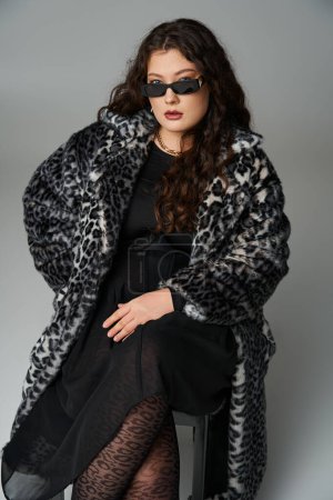 alluring plus size woman in leopard fur coat and sunglasses sitting on black chair with cross legs