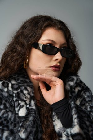 Téléchargez les photos : Portrait de femme courbée glamour en lunettes de soleil regardant du côté avec le menton sur la main sur le gris - en image libre de droit
