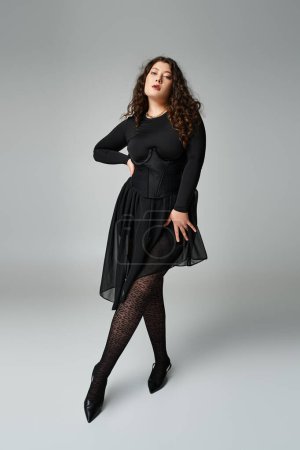 seductora mujer con curvas en traje negro poniendo la pierna hacia adelante con la mano en la cintura sobre fondo gris