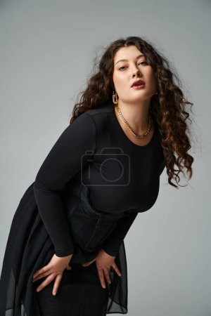 schöne kurvige junge Frau im schwarzen Outfit lehnt sich vor grauem Hintergrund nach vorne