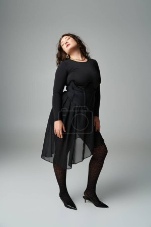lovely curvy woman in black stylish outfit posing with leaning head to behind on grey background