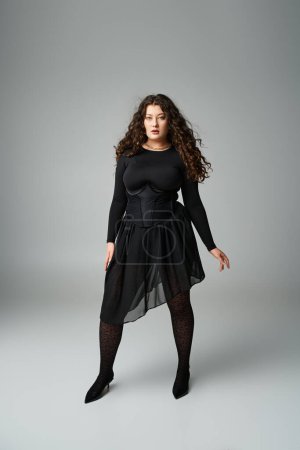 atractiva chica joven con curvas en traje elegante negro de pie con las piernas separadas sobre fondo gris