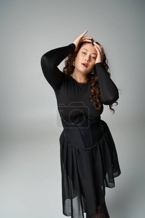 charming curvy young woman in black stylish outfit holding hands to head on grey background