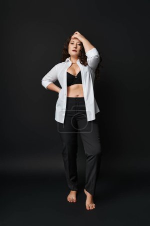 attractive plus size woman in white shirt, black bra and pants with hand on head in dark background