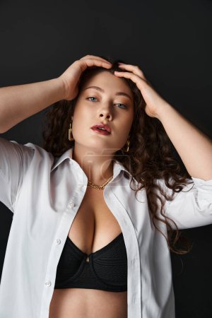 portrait of beautiful plus size woman in white shirt and black bra putting hands on head on grey
