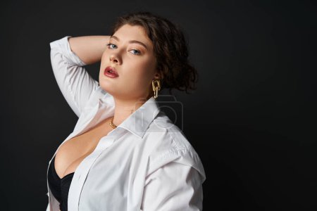 portrait of seductive curvy woman in shirt and bra putting hand behind head and holding hair