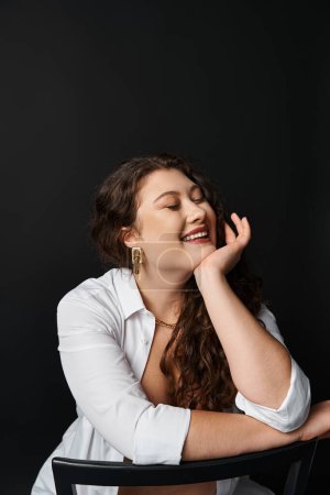 portrait of cheerful plus size woman in her 20s with closed eyes leaning on hand and laughing