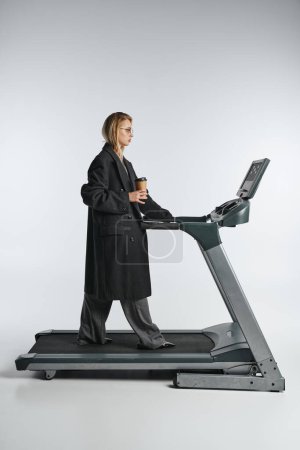 Photo for Appealing graceful woman in stylish black coat with trendy glasses posing on treadmill with coffee - Royalty Free Image