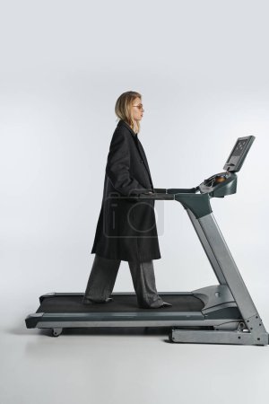 atractiva mujer sofisticada en el abrigo negro de moda posando en la cinta de correr sobre fondo gris