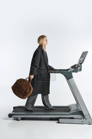 attractive sophisticated woman in fashionable black coat posing on treadmill on gray background