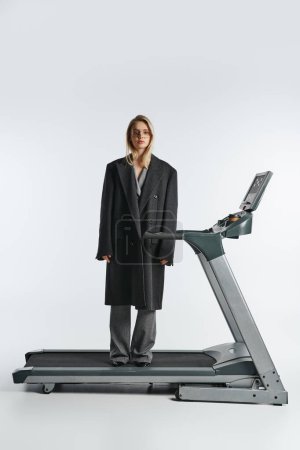 good looking graceful woman in stylish glasses and coat posing on treadmill and looking at camera