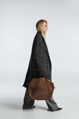 attractive graceful woman with blonde hair in black stylish coat holding brown bag and looking away