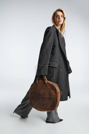 Foto de Atractiva mujer elegante con el pelo rubio en negro elegante abrigo sosteniendo bolsa marrón y mirando hacia otro lado - Imagen libre de derechos