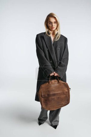 beautiful fashionable woman with glasses and brown bag in black elegant coat looking at camera
