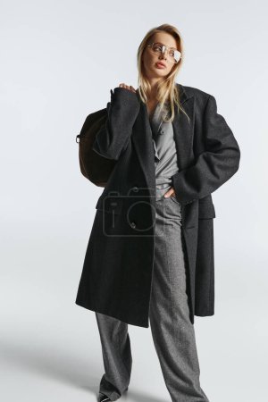 appealing graceful woman with blonde hair in black stylish coat holding brown bag and looking away
