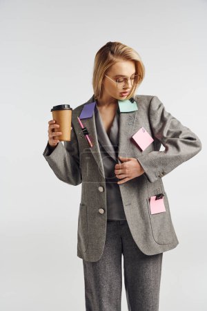 fashionable appealing woman in gray suit with stationery on it posing with coffee and looking away