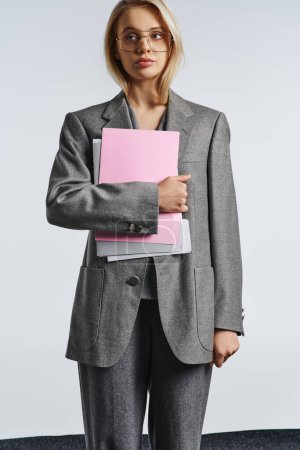 Foto de Atractiva mujer elegante con gafas de estilo en traje gris posando con papeleo y mirando hacia otro lado - Imagen libre de derechos