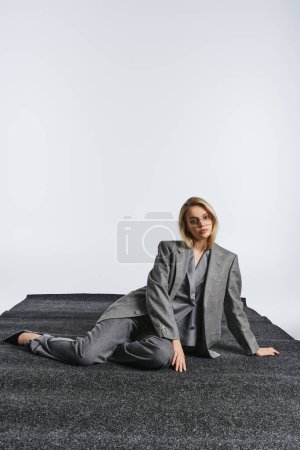 Foto de Mujer elegante de moda con gafas con estilo en traje elegante sentado en el suelo y mirando a la cámara - Imagen libre de derechos