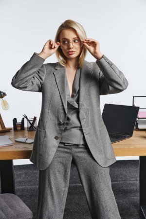 Téléchargez les photos : Belle femme d'affaires élégante en costume intelligent gris posant à côté de la table au bureau et regardant loin - en image libre de droit