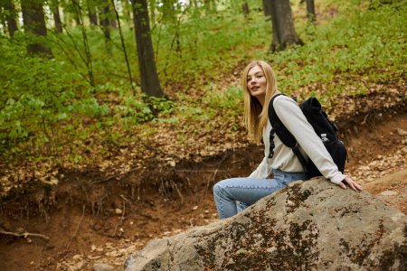 Smiling relaxed blonde woman hiker wearing sweater and backpack sitting on a rock in forest scenery