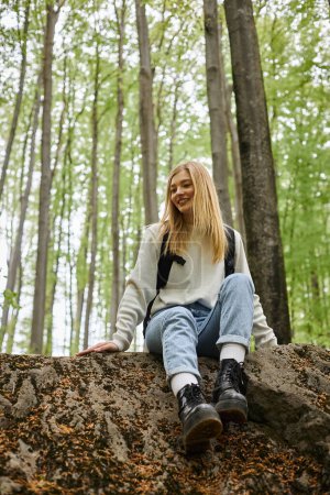 Sonriente senderismo mujer rubia con suéter y mochila sentado en una roca en el bosque