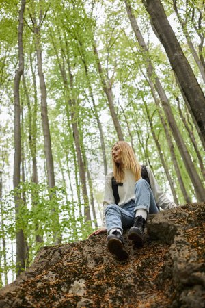 Calm relaxed blond girl hiking in forest sitting on rock and wearing sweater and backpack