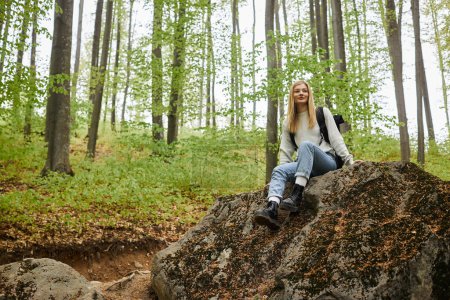Active young blonde woman hiker sitting on a rock on a backpacking trip in peaceful forest