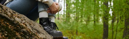 Foto recortada de la mujer excursionista abrazando sus piernas con jeans y botas de senderismo en el bosque, pancarta