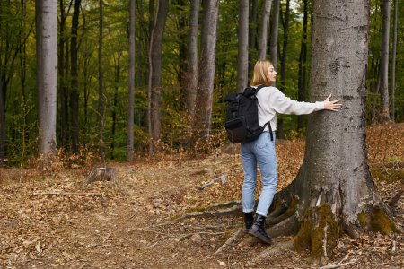 Back view full height portrait of blonde hiker woman in comfy outfit in forest touching a tree