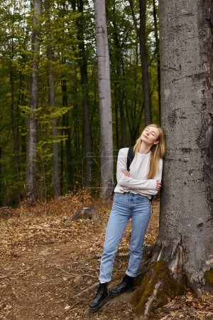 Full height portrait of blonde woman in hiking outfit in forest leaning on tree with eyes closed