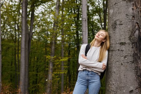 Porträt einer verführerisch entspannten blonden Wanderin im Wald, die sich mit geschlossenen Augen an einen Baum lehnt