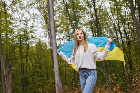 Unterstützende unabhängige blonde Frau steht im Wald und schwenkt die ukrainische Flagge