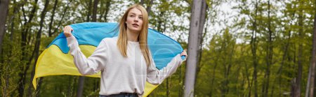 Unterstützende unabhängige blonde Frau, die im Wald steht und die ukrainische Flagge schwenkt, Banner