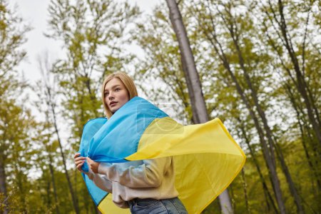 Schöne patriotische blonde Frau steht im Wald und bedeckt Körper mit ukrainischer Flagge