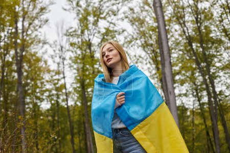 Patriotic woman standing in forest covering body with Ukrainian flag and looking in front of her