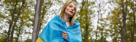 Patriotic woman standing in forest covering body with Ukrainian flag and looking away, banner