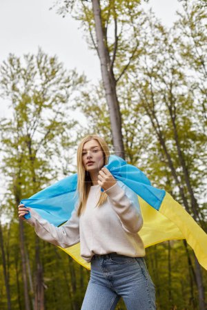 Femme aventurière blonde patriotique tenant fièrement le drapeau national ukrainien dans la forêt, la liberté