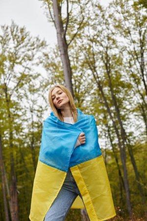 Ernstes blondes Mädchen, das im Wald die ukrainische Flagge über ihrem Körper hält und in die Kamera blickt