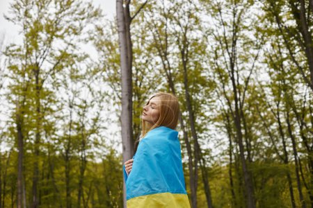 Inspiriertes blondes Mädchen, das im Wald mit geschlossenen Augen die ukrainische Flagge über dem Körper trägt