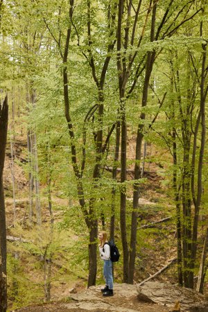 Vista superior de la mujer rubia aventurera en suéter acogedor y jeans con mochila, de pie en el bosque