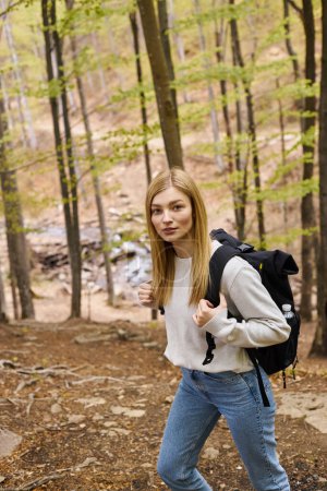 Neugierige blonde Wanderin mit Reiserucksack schlängelt sich durch den Wald und blickt in die Kamera